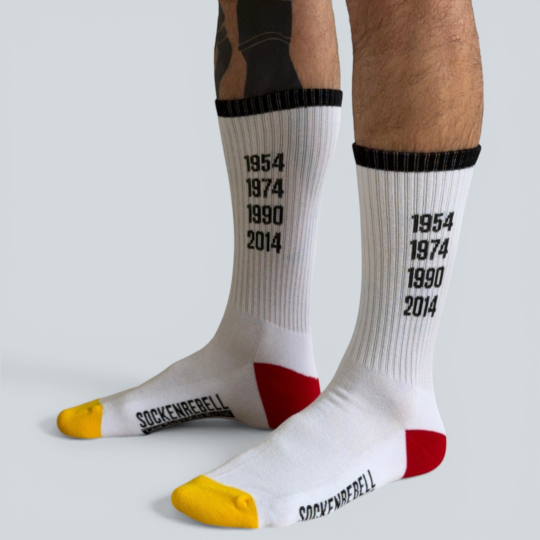 SOCKENREBELL white crew socks with black trim, featuring years 1954, 1974, 1990, 2014 printed vertically, red heel, and yellow toe.